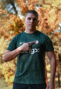 Zaļš sporta krekls ar atkārtojošu "CFC" rakstu, balts "bingx" logos un sarkans un balts lauva emblēma uz kreisā krūšu. Modelis norāda uz emblēmu.