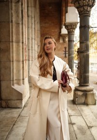 Kremni, prevelik plašč z velikimi žepi in širokimi rokavi, nošen preko temnega zgornjega dela in dolge, lahke krila. Drži bordo torbico.