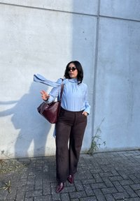 Blusa de manga longa azul-claro, calças castanhas de cintura alta e uma mala bordeaux; textura suave, sapatos de bico e detalhe de lenço fluido.