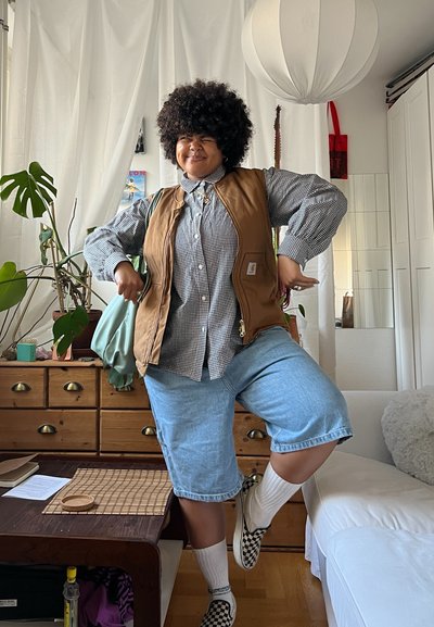 Chaleco marrón con cremallera sobre una camisa de cuadros en blanco y negro, combinado con pantalones cortos de mezclilla azules, calcetines blancos y zapatillas de cuadros sin cordones. Entorno interior.