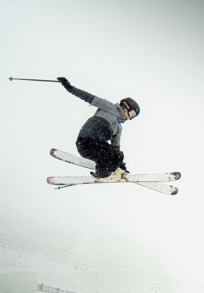 Esquiador con casco negro y chaqueta gris, en el aire con esquís blancos, realizando un salto contra un fondo nevado con suave nieve cayendo.