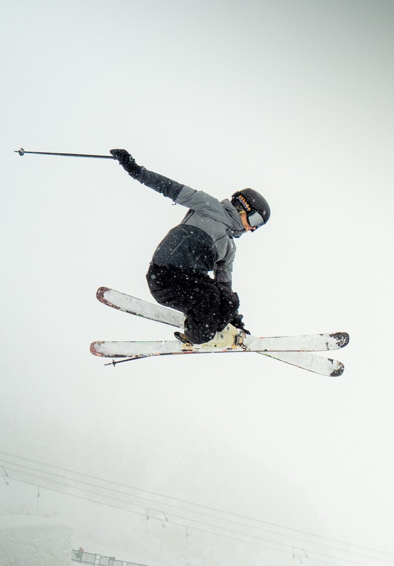 Skieur portant un casque noir et une veste grise, en plein saut avec des skis blancs, contre un décor enneigé avec une chute de neige douce.