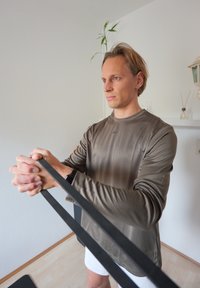 Mężczyzna w brązowej koszuli z długim rękawem używający czarnej taśmy oporowej do ćwiczeń na ramiona w jasnej, minimalistycznej przestrzeni wewnętrznej.