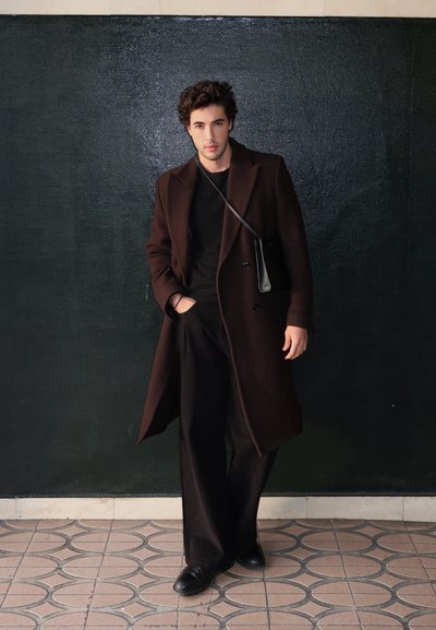 Larga chaqueta marrón sobre una camiseta negra, combinada con pantalones anchos negros y zapatos negros. Un pequeño bolso de hombro negro completa el look.