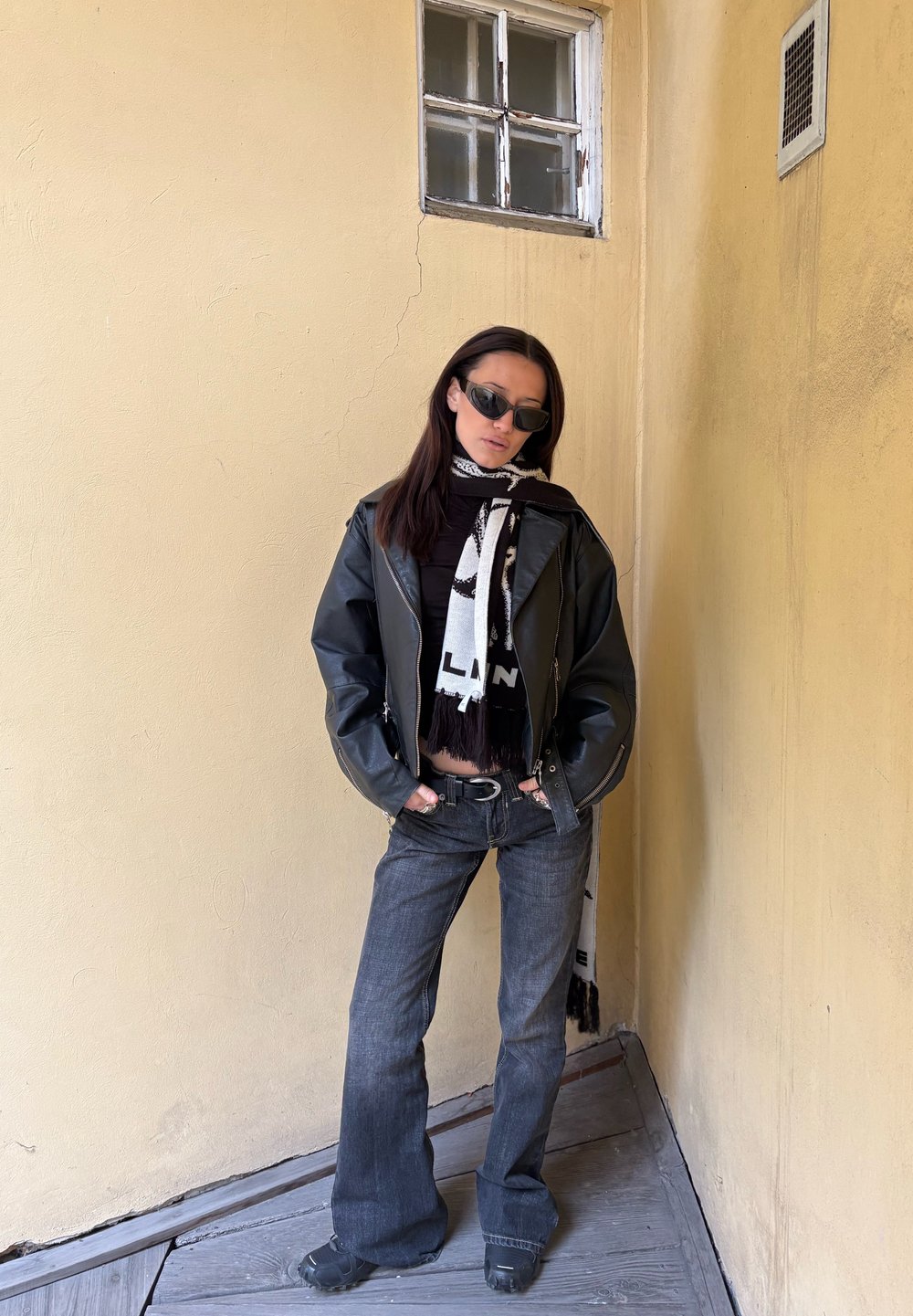 Mustast nahast jakk tumeda pluusi kohal, millele on kombineeritud laienevad tumedad teksad. Kaunistatud must-valge mustriga salliga ja päikeseprillidega.