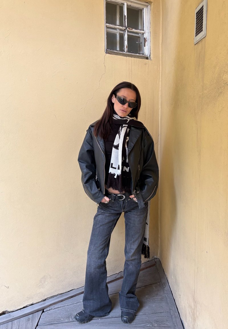 Veste en cuir noire portée par-dessus un haut foncé, associée à un jean évasé foncé. Accessoirisé d'une écharpe noire et blanche à motif et de lunettes de soleil.