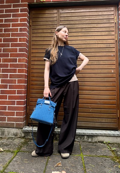 Camiseta azul marino de corte holgado con mangas enrolladas, camiseta interior beige, pantalones de pierna ancha marrón oscuro y un bolso azul acolchado. Fondo de madera y ladrillo.