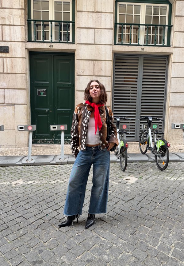 Una mujer con una chaqueta estampada, una bufanda roja y unos vaqueros de pierna ancha está de pie en una calle adoquinada, junto a bicicletas de alquiler y un edificio de piedra.