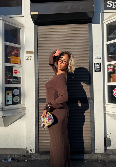 Vestido de punto marrón con canalé, de manga larga y cuello redondo. Sostenido una bolsa texturizada y colorida. De pie frente a una ventana con persianas.