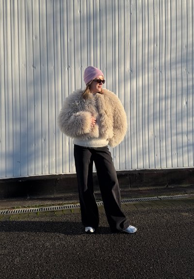 Persona con chaqueta beige peluda, gorro rosa, gafas de sol, pantalones negros y zapatos blancos de pie sobre el pavimento junto a una pared de metal corrugado.