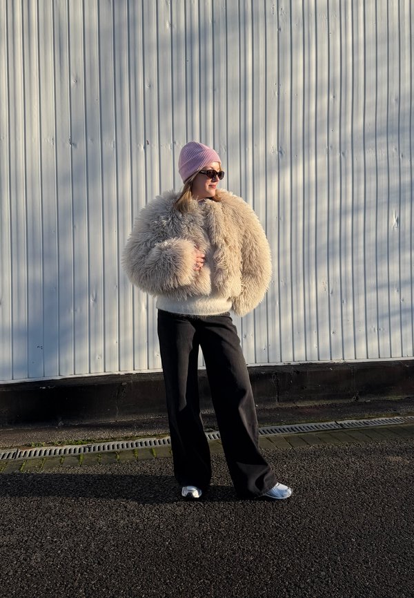 Persona con giacca beige pelosa, berretto rosa, occhiali da sole, pantaloni neri e scarpe bianche in piedi sul marciapiede accanto a un muro di lamiera ondulata.