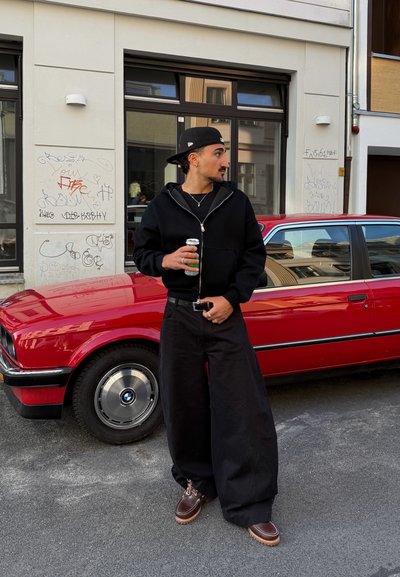 Una persona en una sudadera con cremallera negra y pantalones anchos negros está de pie al lado de un coche rojo, sosteniendo una lata. La escena presenta arte urbano de fondo.
