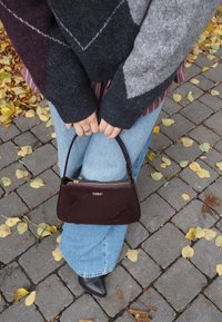 Burgundská lesklá kabelka zaobleného tvaru so zipsovým uzáverom a zlatým logom. V kombinácii s svetlomodrými džínsami a vzorovaným šedým sveterom.