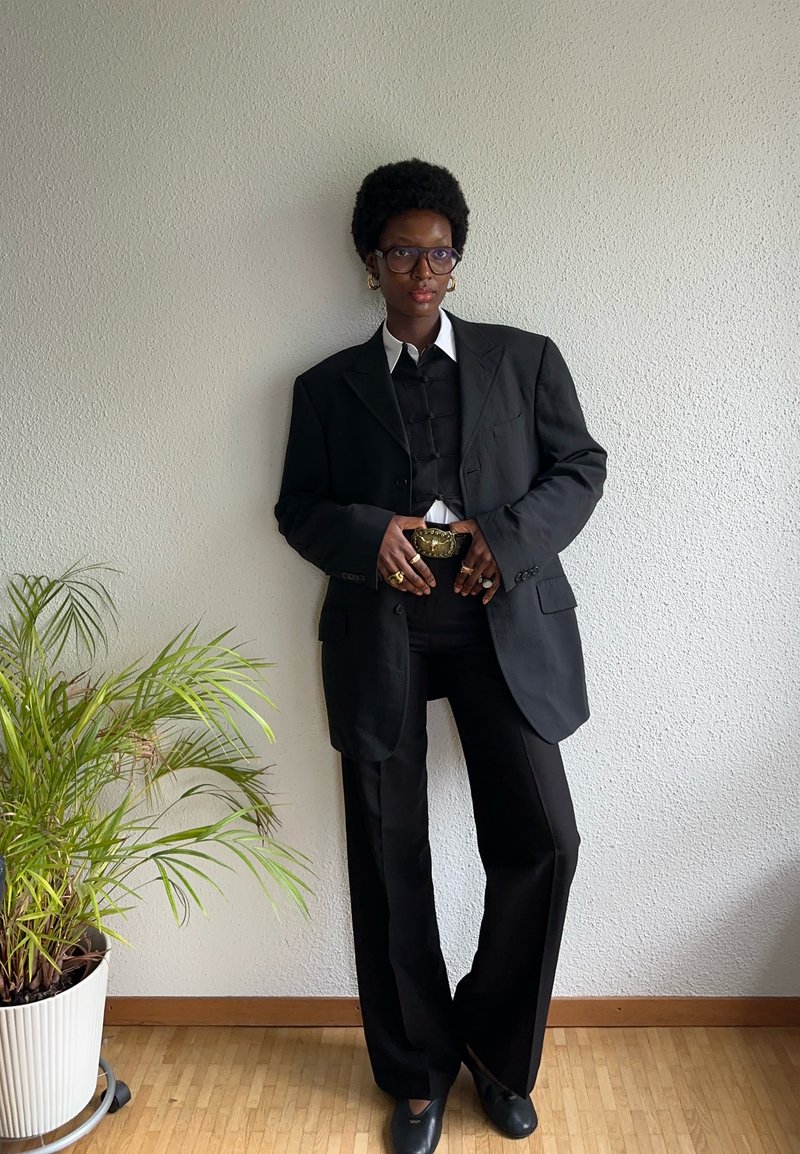 Persona con capelli corti e ricci e occhiali, che indossa un blazer oversize nero, pantaloni neri, una camicia bianca e una cintura decorativa, in piedi all'interno vicino a una pianta.