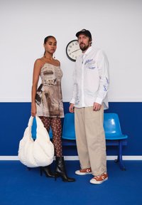 Two models stand in front of a clock. The woman wears a patterned beige dress, brown leggings, and black heeled boots. The man wears a white shirt and beige trousers with orange trainers. Both are holding a puffy white bag.