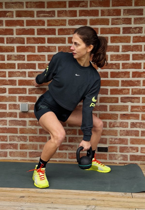 Kvinne i treningstøy som gjør knebøy på en matte, holder en kettlebell i den ene hånden foran en murvegg.