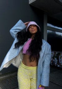 Svetlomodrý vlnený kabát, ružový vedro klobúk, fialový skrátený sveter a štruktúrované žlté nohavice. Dlhé tmavé vlasy, drží klobúk s dôvtipným postojom.