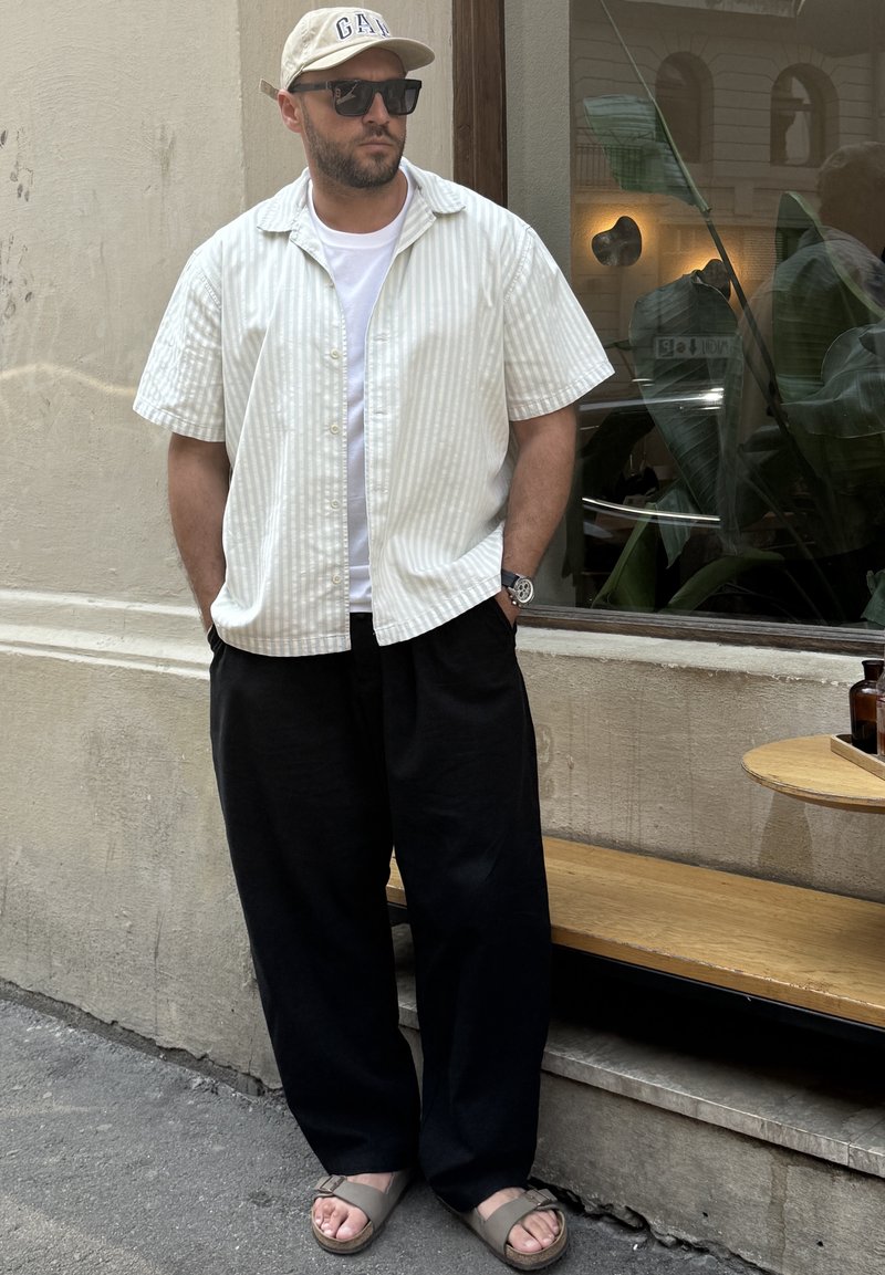Korte mouwen, gestreept wit shirt over een effen wit t-shirt; zwarte wijde broek; beige sandalen; pet; zonnebril; staand bij een raam.