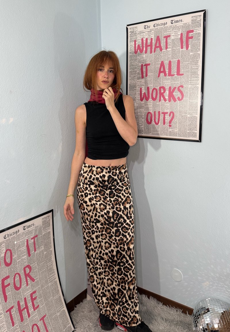 Giovane donna con top nero senza maniche e gonna con stampa leopardata in piedi accanto a due poster con testo rosso su una parete chiara uniforme e una palla da discoteca.