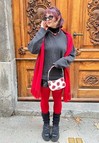 Maglione grigio oversize, sciarpa rossa con frange, gonna nera, collant rossi, calze nere morbide, stivali neri chunky e una borsa bianca con stampa di fragole.