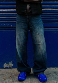 Φαρδιά μπλε τζιν με πλυμένα σημεία, φορεμένα με bold μπλε αθλητικά παπούτσια που διαθέτουν υφές. Το φόντο είναι ένας ενιαίος μπλε τοίχος.