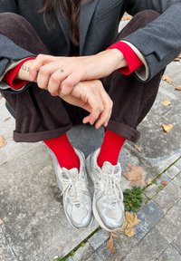 Ασημένια αθλητικά παπούτσια, κόκκινες ριγέ κάλτσες, καφέ παντελόνι από καρό ύφασμα, γκρι σακάκι. Ορατά τατουάζ στα χέρια και στους καρπούς.