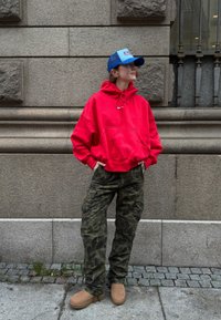 Sudadera roja con capucha, bolsillo frontal y logo, pantalones cargo verdes con camuflaje, botas beige sin cordones y una gorra azul. Fondo urbano.