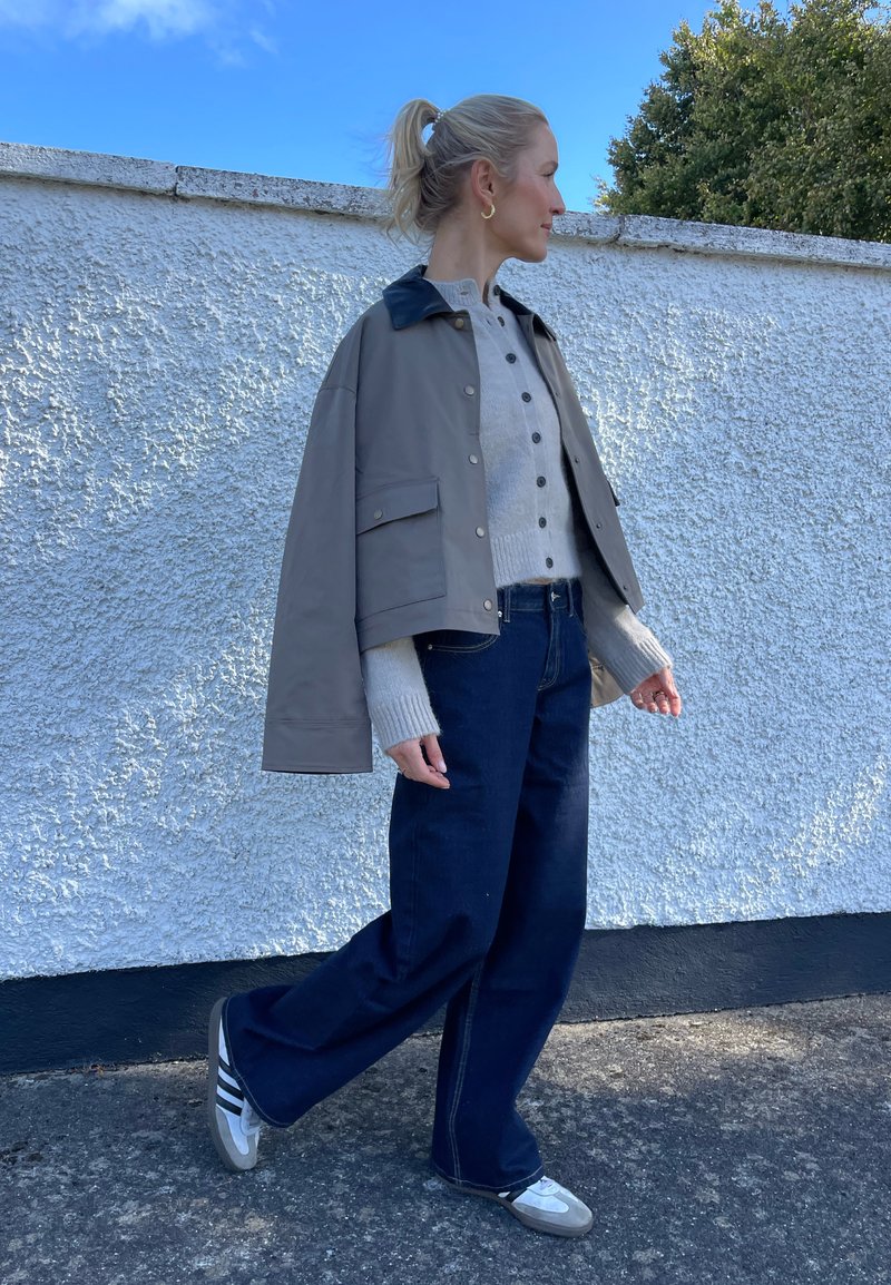 Hellgraue Cropped-Jacke mit dunklem Kragen, beiger Strickpullover, marineblaue weite Jeans und graue Sneaker mit schwarzen Streifen.