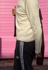 Beige geribd shirt met lange mouwen en tribale print, gecombineerd met zwarte joggers met luipaardprint, witte strepen en Adidas-logo.