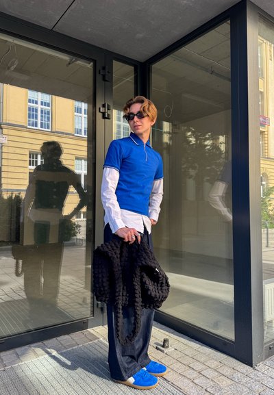 Camisa polo azul con cremallera, camisa blanca por debajo, pantalones anchos oscuros, bolsa negra con textura y zapatillas azules. Reflejo visible en la puerta de cristal.