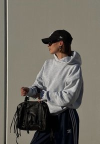 Sweatshirt cinza com capuz, boné preto, óculos de sol e grande bolsa de couro preta com franjas. Calças escuras com riscas laterais e um design simples.