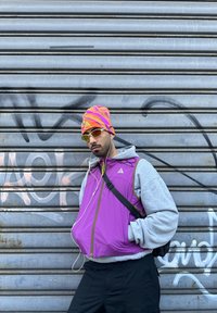 Homme portant un gilet violet, un sweat à capuche gris, un bonnet coloré et des lunettes de soleil, écoutant des écouteurs, debout devant un rideau métallique tagué.