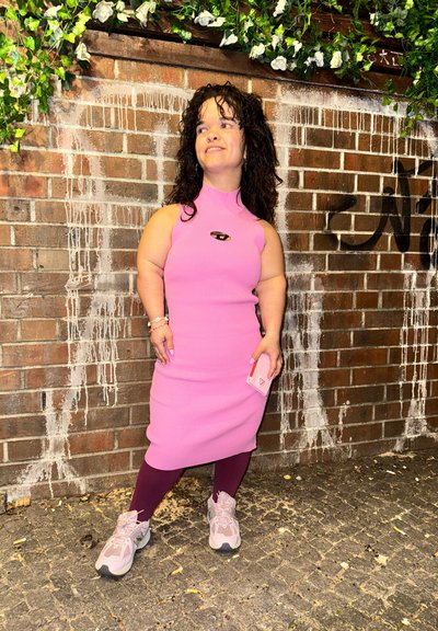 Vestido rosa de canalé con cuello alto, combinado con leggings burdeos y zapatillas rosa claro. Pequeña cartera rosa sostenida en la mano derecha. Fondo de ladrillo.
