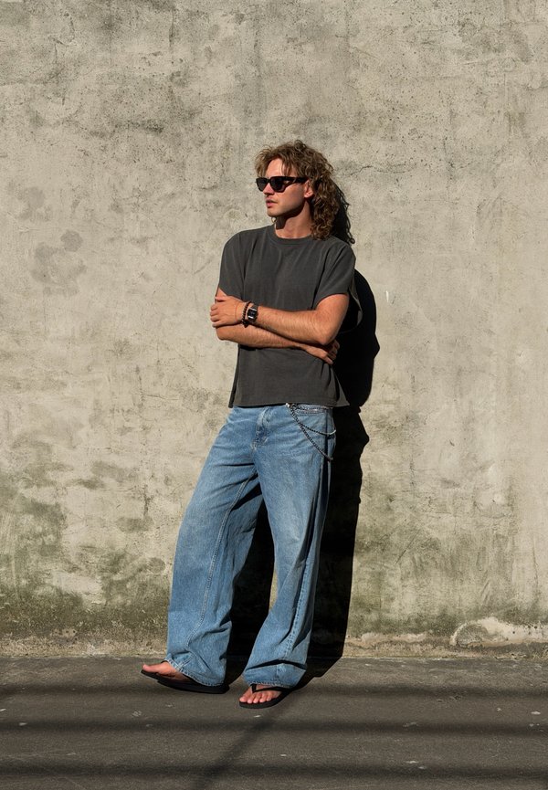 Mann mit lockigem Haar und Sonnenbrille steht barfuß vor einer strukturierten Betonwand, trägt ein dunkles Hemd und lockere blaue Jeans mit Kettenaccessoire.