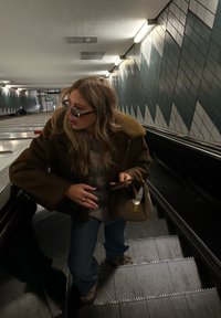 Smeđa oversized jakna s krznеним ovratnikom, svijetlo sivi pulover, plave traperice, drži telefon i smeđu torbicu dok se nalazi na pokretnim stepenicama.