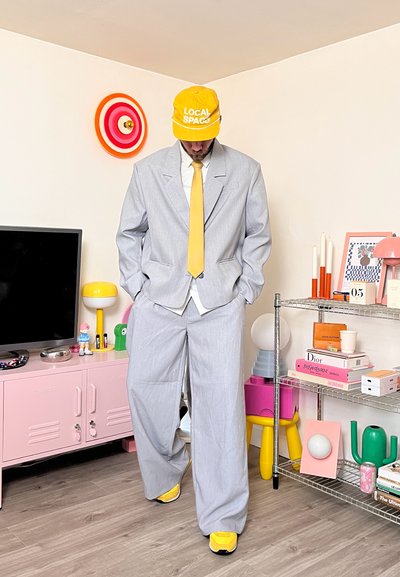 Hombre con traje gris claro de gran tamaño, corbata amarilla, gorra amarilla y zapatos amarillos, de pie en una habitación colorida con estanterías y objetos decorativos.