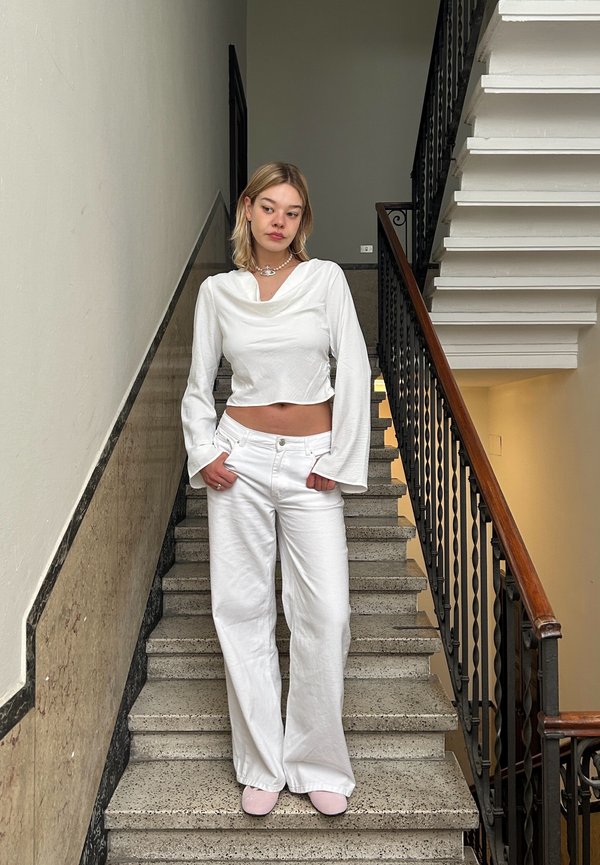 Top bianco a maniche lunghe e corte con scollatura drappeggiata, abbinato a pantaloni bianchi larghi. In piedi su una scala con un corrimano in un ambiente neutro.