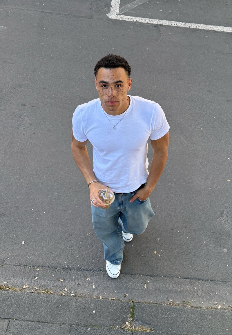 Jeune homme avec taches de rousseur portant un t-shirt blanc et un jean tient une boisson glacée, marchant sur l’asphalte près des lignes de parking marquées, regardant vers le haut.
