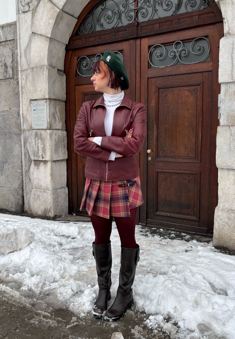 Veste en cuir marron, col roulé blanc, jupe à carreaux rouge, jaune et bleu, leggings bordeaux et bottes noires montantes jusqu'aux genoux. Sol enneigé.