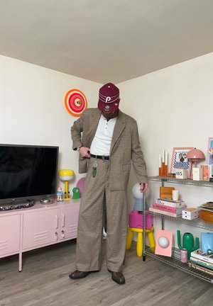 Persona vestida con un traje marrón de cuadros, camisa blanca y gorra granate en una habitación decorada con estanterías coloridas, libros y un televisor.