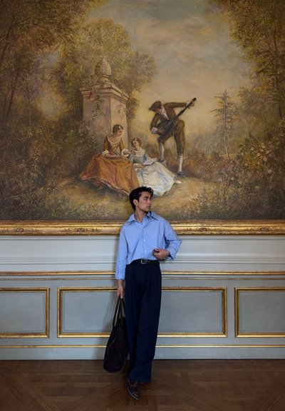 Un hombre con una camisa azul claro y pantalones oscuros se encuentra frente a un mural que representa a dos mujeres con vestidos y a un hombre tocando un instrumento de cuerda.