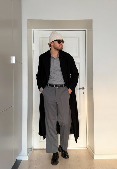 Hombre con gorro de punto blanco, gafas de sol negras, polo gris, abrigo largo negro, pantalones grises y zapatos negros de pie en interior con las manos en los bolsillos.