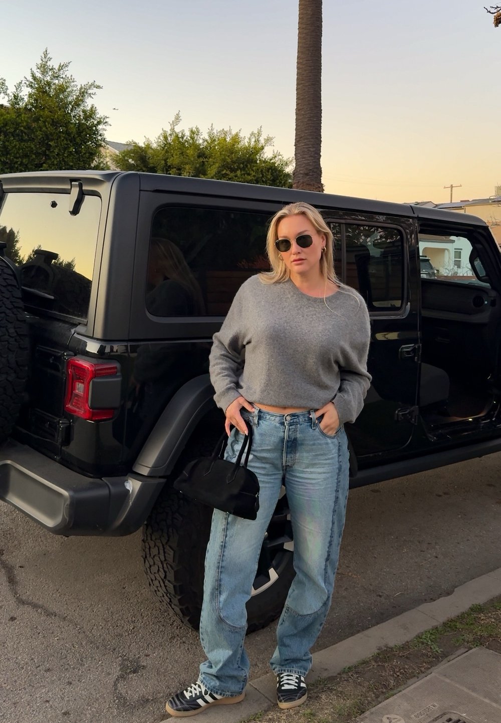 Femme portant des lunettes de soleil, un pull gris et un jean, debout, tenant un sac à main noir à côté d'un véhicule tout-terrain noir au coucher du soleil.