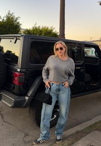 Femme portant des lunettes de soleil, un pull gris et un jean, debout, tenant un sac à main noir à côté d'un véhicule tout-terrain noir au coucher du soleil.