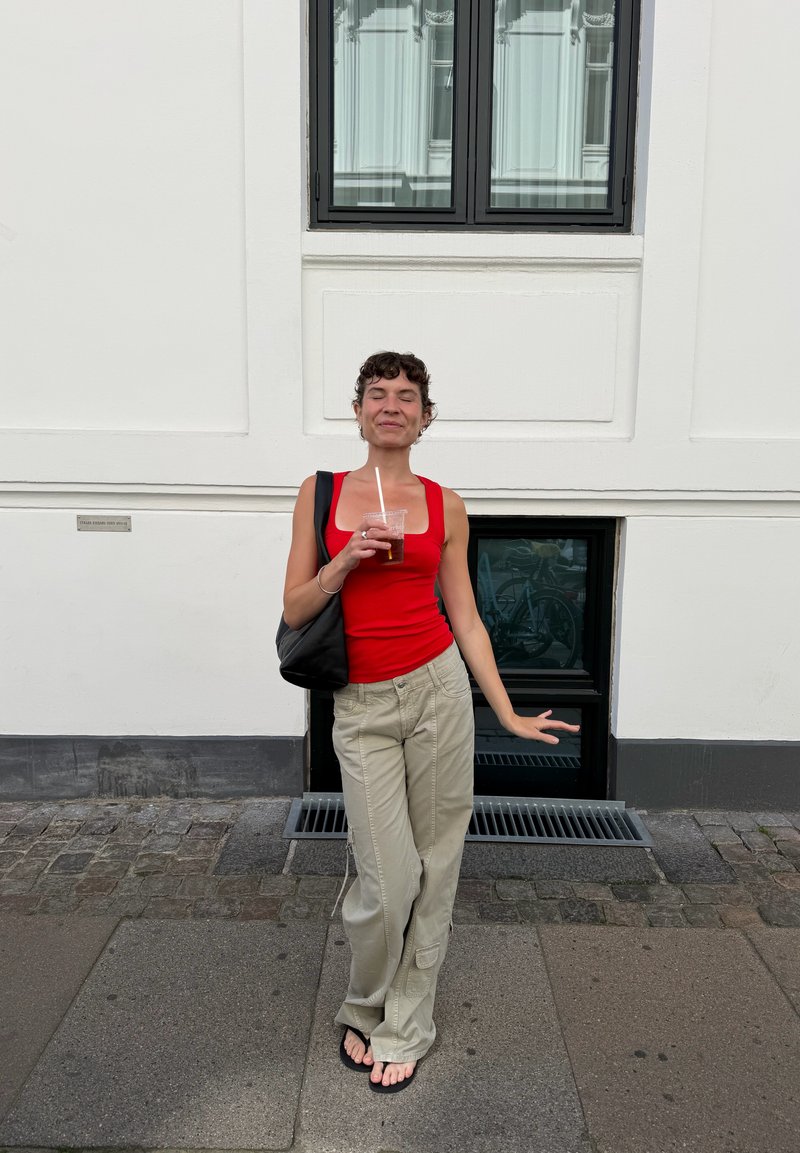 Femme aux cheveux courts et bouclés portant un débardeur rouge et un pantalon beige, debout sur le trottoir, tenant une boisson glacée les yeux fermés et un sac noir sur l'épaule.