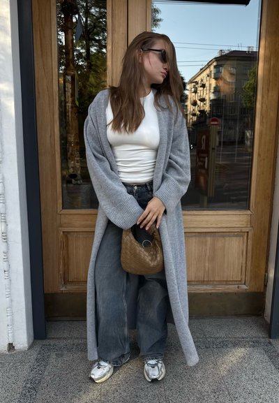 Cárdigan de punto gris sobre una blusa blanca, combinado con jeans oscuros holgados y zapatillas metálicas. Sosteniendo un bolso de mimbre marrón. Fondo de madera.