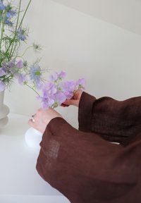 Λευκό βάζο που κρατάει μοβ και μπλε λουλούδια, με λεπτά πράσινα κοτσάνια. Ορατοί καφέ λινά μανίκια καθώς τα χέρια τακτοποιούν τα λουλούδια.