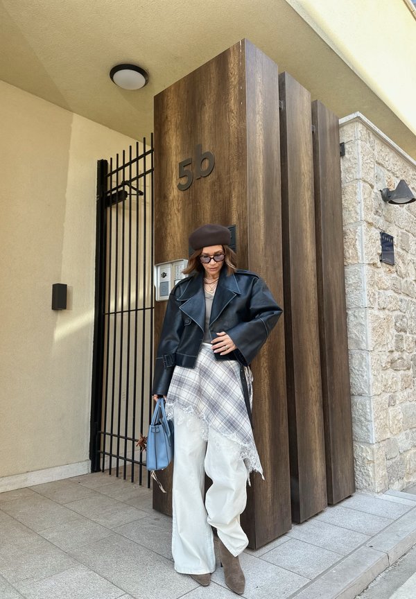Vrouw draagt een zwarte leren jas, geruite rok over witte broek, bruin baret, zonnebril, houdt een blauwe handtas vast, staat bij een houten deur met nummer 5b.