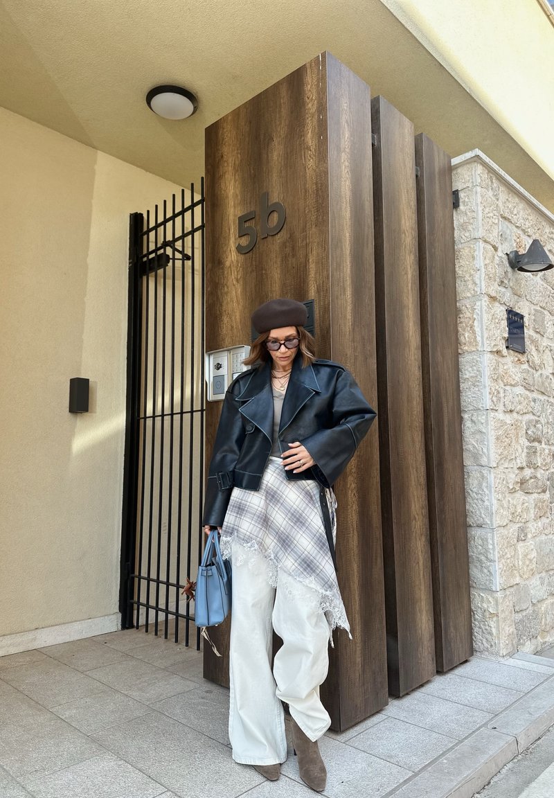 Γυναίκα με μαύρο δερμάτινο μπουφάν, επικάλυψη φούστας καρό, λευκό παντελόνι, καφέ μπερέ, γυαλιά ηλίου, κρατώντας μπλε τσάντα, στέκεται δίπλα σε ξύλινη πόρτα με αριθμό 5β.