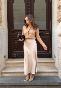 Bež kardigan od vune s gumbima, krem saten suknja do poda i smeđi gležnjači. Drži malu crnu torbicu i stoji na kamenim stepenicama.